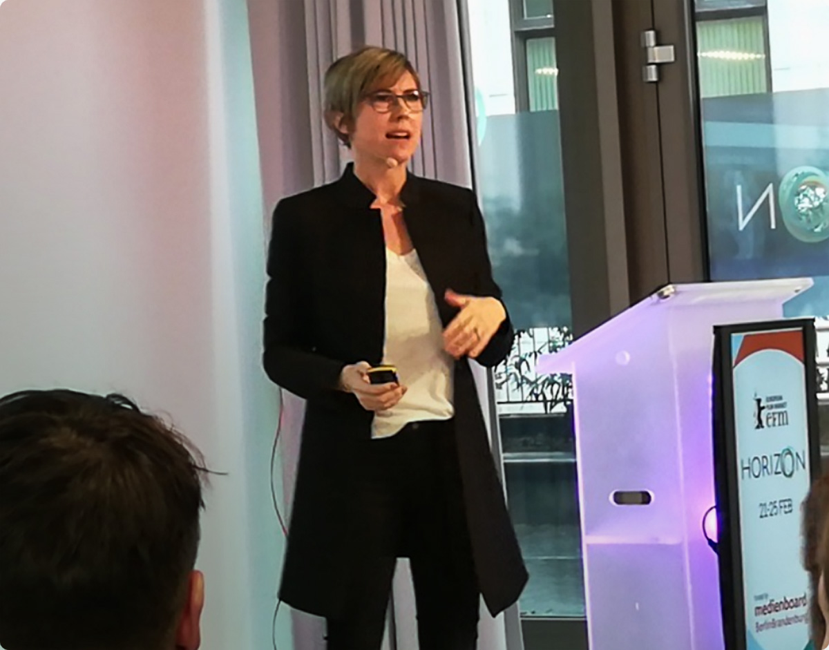 Monica Landers stands onstage at a speaking event, wearing a microphone and black jacket